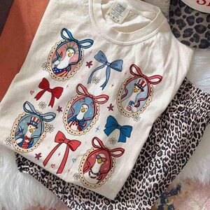 Op de afbeelding: Cr&egrave;me Comfort Colors T-shirt met een patriottisch ganzenontwerp in ovale frames, rode, witte en blauwe strikken. Het shirt wordt gecombineerd met een pyjamabroek met luipaardprint. Het shirt heeft de tekst "Comfort Colors".