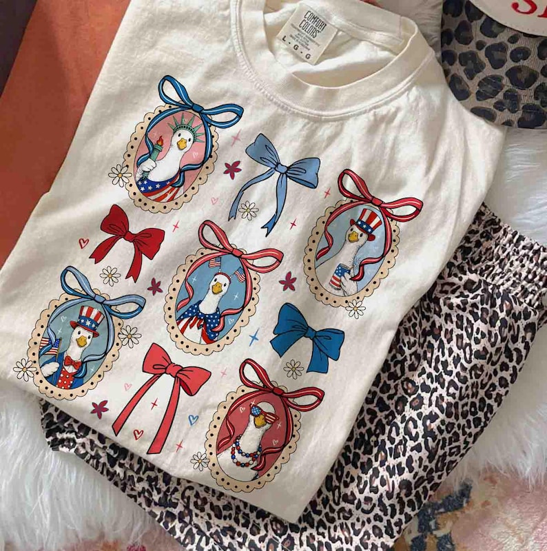 Op de afbeelding: Cr&egrave;me Comfort Colors T-shirt met een patriottisch ganzenontwerp in ovale frames, rode, witte en blauwe strikken. Het shirt wordt gecombineerd met een pyjamabroek met luipaardprint. Het shirt heeft de tekst "Comfort Colors".