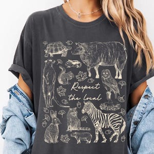 May include: Dark gray t-shirt featuring a collection of sketched animals including a zebra, owl, and meerkat. The phrase "Respect the Local" is printed in a script font. The shirt has short sleeves and a relaxed fit.