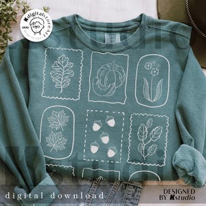 May include: Teal sweatshirt featuring white line art of fall elements like leaves, pumpkins, and acorns within decorative frames. The words "digital download" and "designed by Kstudio" are printed on the bottom of the sweatshirt.