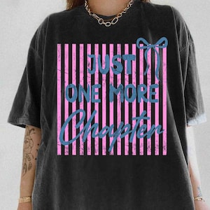 Puede incluir: Camiseta negra con un gr&aacute;fico desgastado. El gr&aacute;fico presenta rayas verticales rosas, texto azul que dice "JUST ONE MORE CHAPTER" y un lazo azul. La camiseta tiene mangas cortas y un corte holgado.