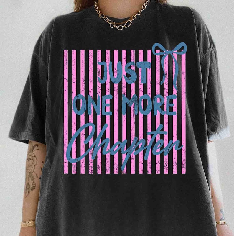 Puede incluir: Camiseta negra con un gr&aacute;fico desgastado. El gr&aacute;fico presenta rayas verticales rosas, texto azul que dice "JUST ONE MORE CHAPTER" y un lazo azul. La camiseta tiene mangas cortas y un corte holgado.