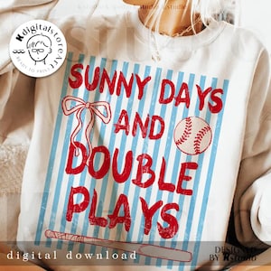 May include: Cream-coloured sweatshirt with a baseball-themed design. The graphic includes the text "SUNNY DAYS AND DOUBLE PLAYS" in red, a baseball, a bow, and a bat. The words "digital download" are also visible.