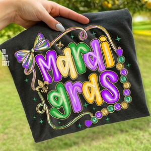 May include: Black t-shirt with a colorful "Mardi Gras" design. The design features the text in purple, green, and yellow, with a bow, fleur-de-lis, and bead accents. The shirt is held by a hand.