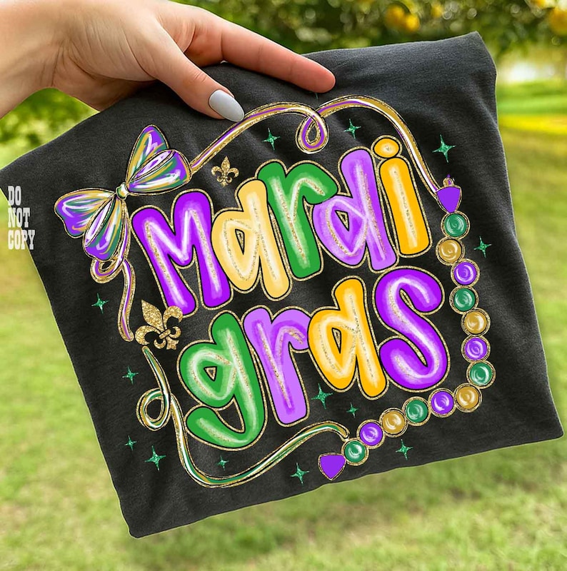 May include: Black t-shirt with a colorful "Mardi Gras" design. The design features the text in purple, green, and yellow, with a bow, fleur-de-lis, and bead accents. The shirt is held by a hand.