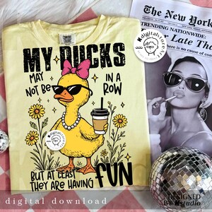 Puede incluir: Camiseta amarillo claro con un gr&aacute;fico de un pato amarillo con gafas de sol, un lazo rosa y un collar de perlas. El texto dice: "MY DUCKS MAY NOT BE IN A ROW BUT AT LEAST THEY ARE HAVING FUN."