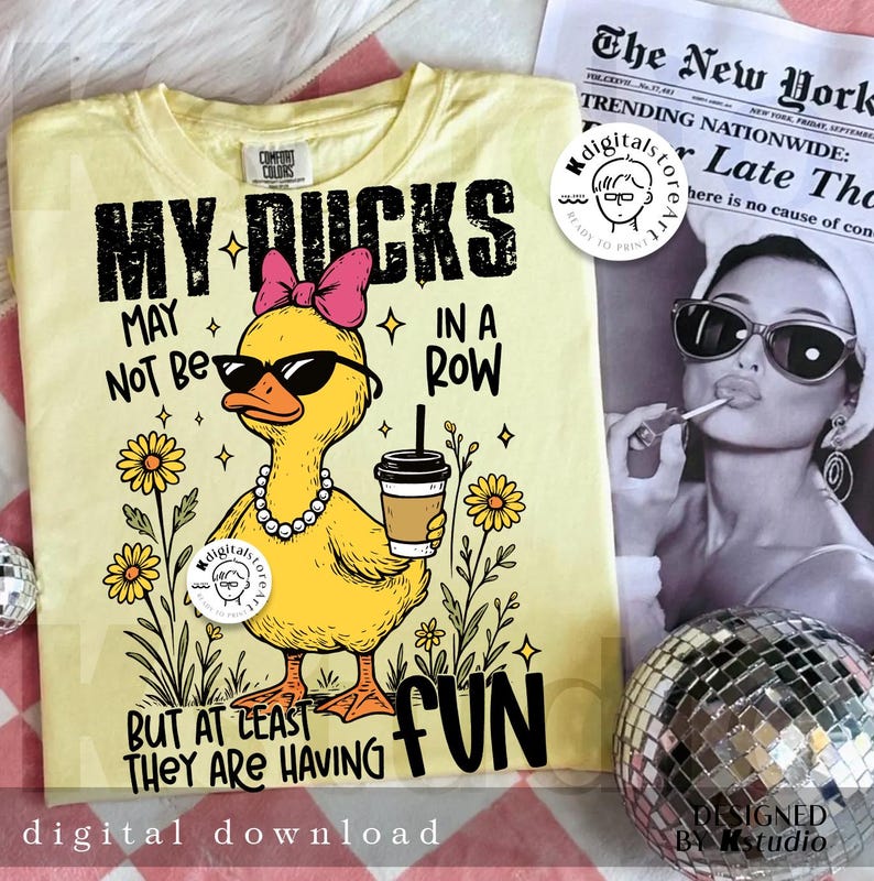 Puede incluir: Camiseta amarillo claro con un gr&aacute;fico de un pato amarillo con gafas de sol, un lazo rosa y un collar de perlas. El texto dice: "MY DUCKS MAY NOT BE IN A ROW BUT AT LEAST THEY ARE HAVING FUN."