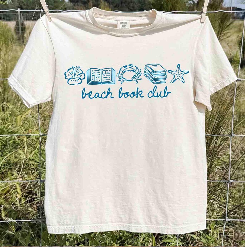 May include: Cream-colored t-shirt with a blue graphic of a flower, book, crab, stacked books, and starfish. The words "beach book club" are printed in blue below the graphic. The shirt is hanging on a clothesline.