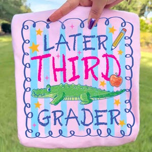 Op de afbeelding: Roze t-shirt met de woorden "Later Third Grader" in blauw en roze. Het ontwerp bevat een groene alligator, een gele potlood, een oranje appel en gele sterren. Het t-shirt wordt door een persoon vastgehouden.