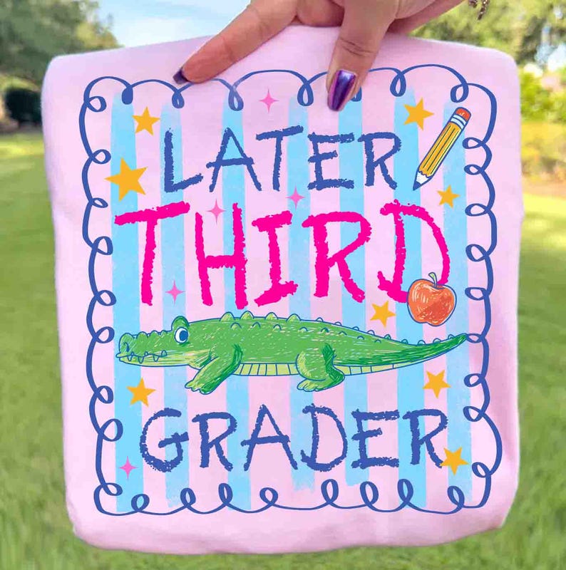 Op de afbeelding: Roze t-shirt met de woorden "Later Third Grader" in blauw en roze. Het ontwerp bevat een groene alligator, een gele potlood, een oranje appel en gele sterren. Het t-shirt wordt door een persoon vastgehouden.