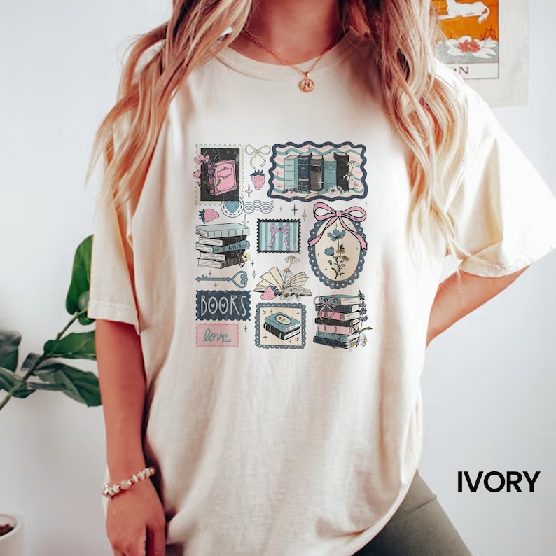 May include: Ivory-colored t-shirt featuring a collage of book-themed illustrations. The design includes stacks of books, stamps, and floral elements in shades of blue, pink, and green. The word "BOOKS" and "love" are also included in the design.