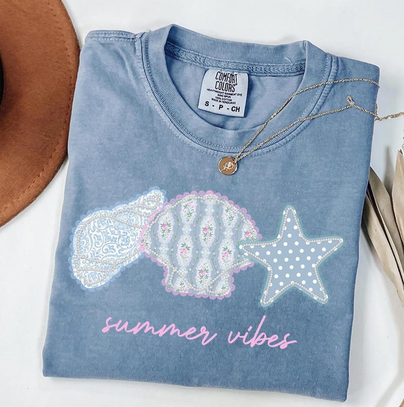May include: A blue Comfort Colors t-shirt with a seashell, paisley, and starfish design. The shirt has the words "summer vibes" in pink script. A gold necklace with a pendant is on the shirt.