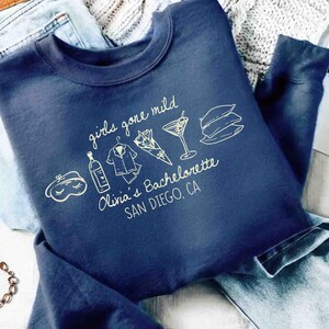 May include: Navy blue sweatshirt with the text "girls gone mild" and "Olivia's Bachelorette SAN DIEGO, CA" in white script. The design includes illustrations of a sleep mask, bottle, pajamas, bouquet, cocktail, and pillows.