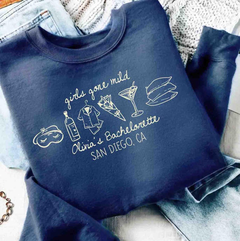 May include: Navy blue sweatshirt with the text "girls gone mild" and "Olivia's Bachelorette SAN DIEGO, CA" in white script. The design includes illustrations of a sleep mask, bottle, pajamas, bouquet, cocktail, and pillows.