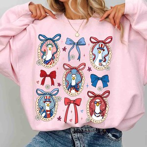 Op de afbeelding: Roze sweatshirt met een herhaald patroon van cartoon ganzen in patriottische outfits, omlijst door decoratieve strikken en bloemaccenten. De ganzen zijn afgebeeld met Amerikaanse vlagmotieven en accessoires.