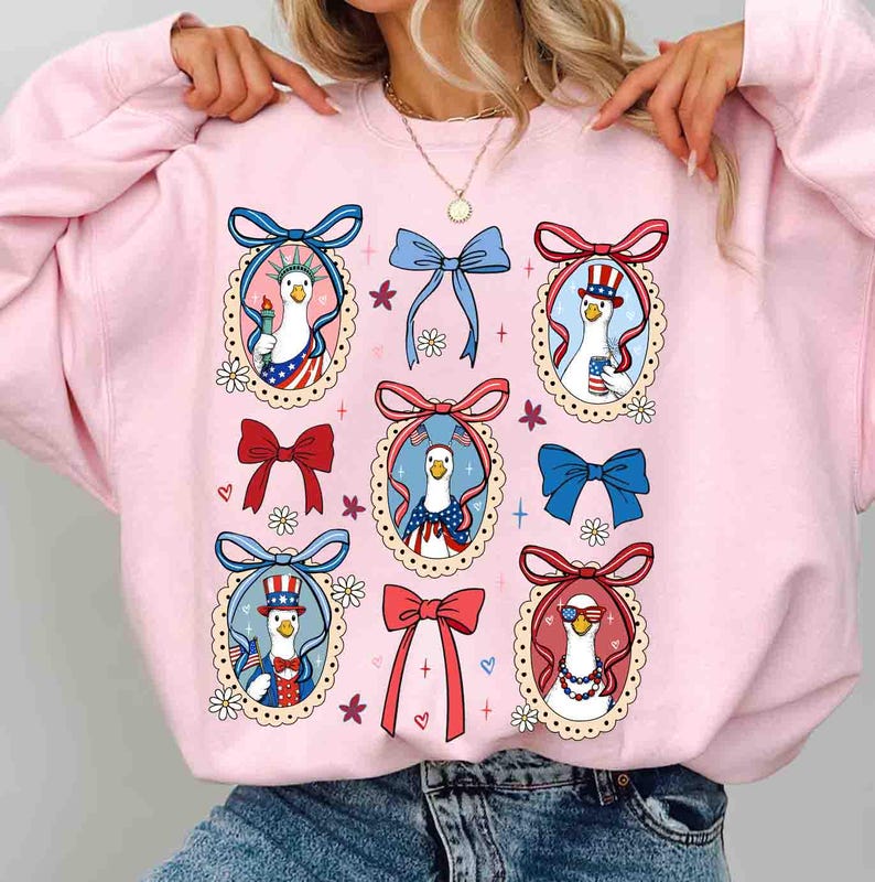 Op de afbeelding: Roze sweatshirt met een herhaald patroon van cartoon ganzen in patriottische outfits, omlijst door decoratieve strikken en bloemaccenten. De ganzen zijn afgebeeld met Amerikaanse vlagmotieven en accessoires.