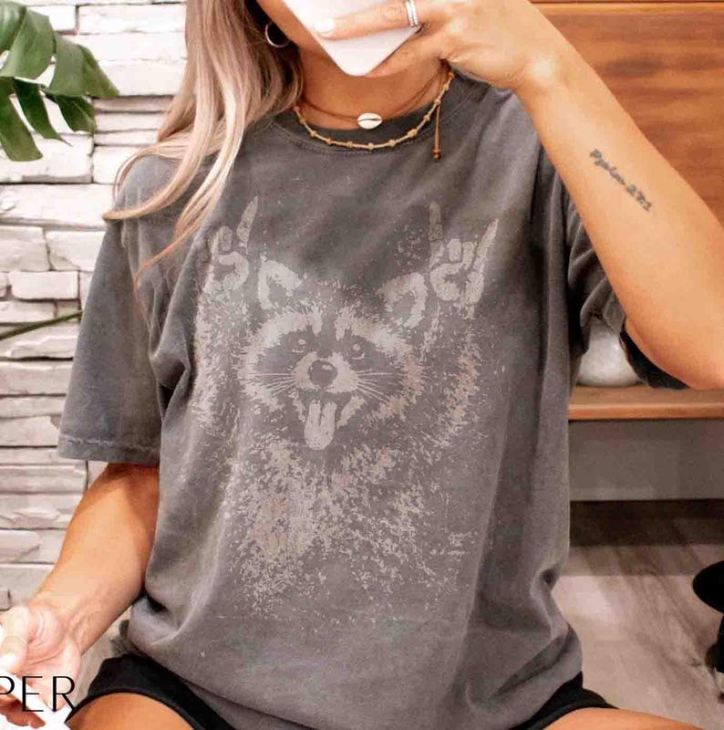 Pu&ograve; includere: T-shirt grigio con grafica di un procione che fa il gesto rock and roll. La lingua del procione &egrave; visibile. La maglietta ha un aspetto vintage e usurato.