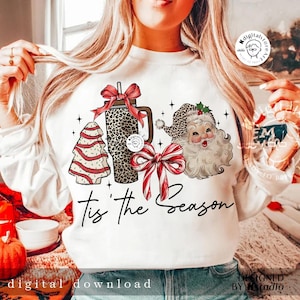 May include: White sweatshirt with a Christmas design featuring a leopard print tumbler, a Christmas tree, a Santa Claus, and a red and white bow. The text "Tis the Season" is written in black script.