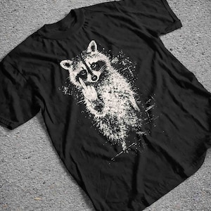 May include: Black t-shirt featuring a white graphic of a raccoon giving the middle finger. The raccoon is depicted in a distressed, splattered style. The t-shirt has a crew neck and short sleeves.