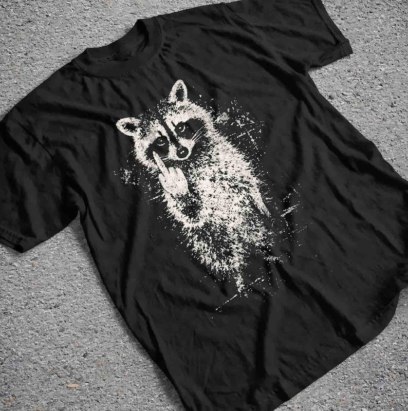 May include: Black t-shirt featuring a white graphic of a raccoon giving the middle finger. The raccoon is depicted in a distressed, splattered style. The t-shirt has a crew neck and short sleeves.
