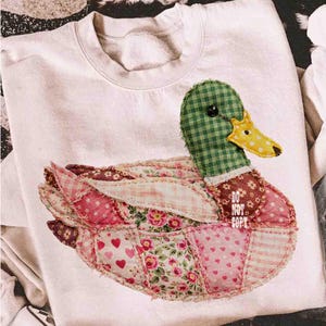 May include: White sweatshirt featuring a patchwork duck design. The duck has a green checkered head, a yellow beak, and wings made of various pink and floral patterned fabrics. The text "DO NOT COPY" is visible on the duck's body.