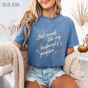 May include: Blue jean colored t-shirt with the text "That sounds like my husband's problem" in a cursive font. The design includes leopard print bows and star accents. The shirt is a casual style.