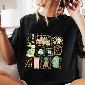 May include: Black t-shirt featuring a colorful Easter-themed graphic. The design includes framed illustrations of Easter eggs, bunnies, teacups, and bows. The artwork is in a vintage, floral style, with pastel colors and decorative borders.