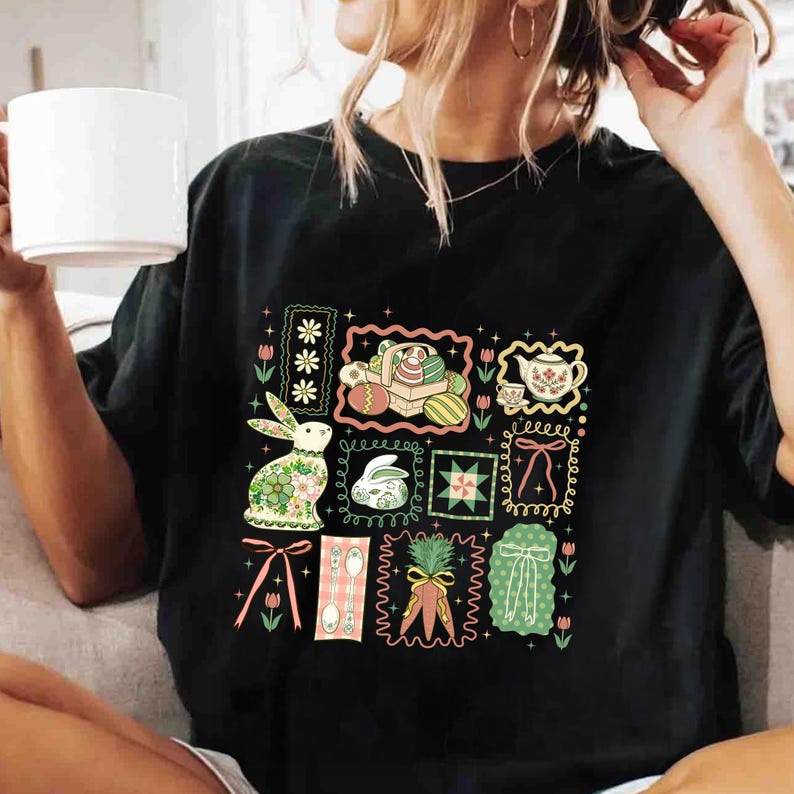 May include: Black t-shirt featuring a colorful Easter-themed graphic. The design includes framed illustrations of Easter eggs, bunnies, teacups, and bows. The artwork is in a vintage, floral style, with pastel colors and decorative borders.