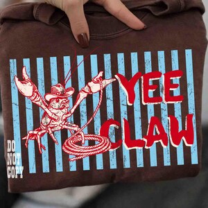 May include: Brown sweatshirt with a distressed graphic featuring a cowboy crawfish with a rope and the words "YEE CLAW" in red. Blue vertical stripes are behind the crawfish. The words "DO NOT COPY" are in the lower left corner.