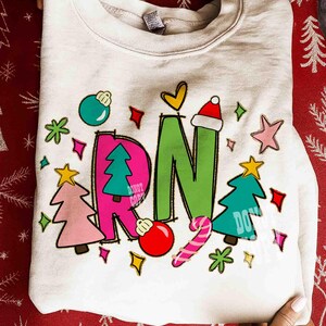May include: Cream-colored sweatshirt with a festive design. The design includes the letters "RN" in green and pink, decorated with Christmas ornaments, trees, stars, and a candy cane. A Santa hat tops the letter "N".