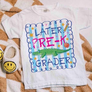 Op de afbeelding: Wit T-shirt met een kleurrijke afbeelding met de tekst "Later Pre-K Grader". Het ontwerp bevat een groene alligator, een rode appel, een gele potlood en blauwe en gele sterren. Het T-shirt is voor kinderen.