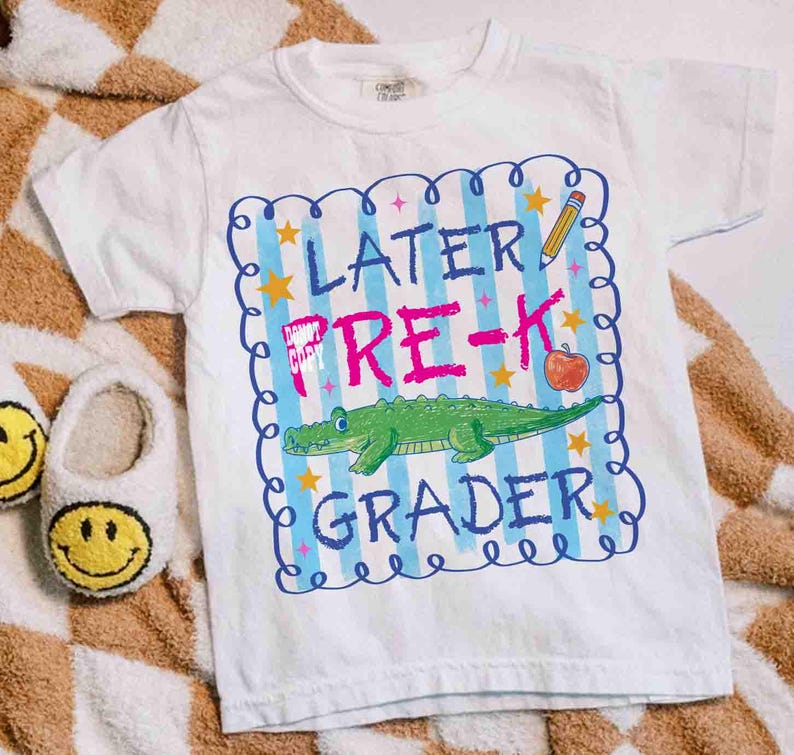 Op de afbeelding: Wit T-shirt met een kleurrijke afbeelding met de tekst "Later Pre-K Grader". Het ontwerp bevat een groene alligator, een rode appel, een gele potlood en blauwe en gele sterren. Het T-shirt is voor kinderen.