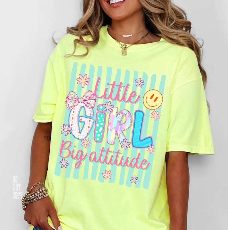May include: A neon yellow short-sleeve t-shirt featuring a graphic print with light blue vertical stripes and the text Little Girl Big attitude in pink and blue. The design includes a patterned bow, small flowers, and a yellow smiley face.