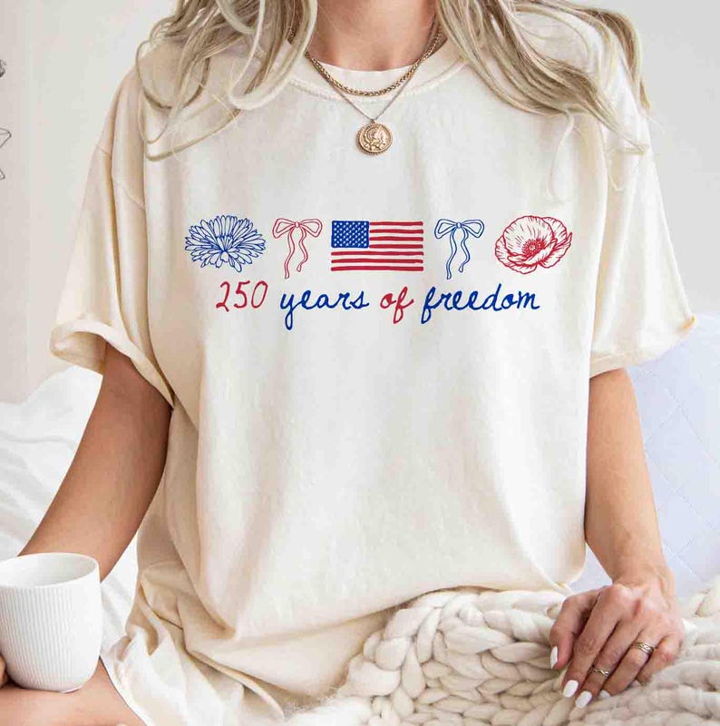 May include: Cream-colored t-shirt with a patriotic design. The shirt features a blue flower, two red bows, an American flag, and a red poppy. The text "250 years of freedom" is written in blue and red.