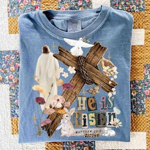 May include: A light blue Comfort Colors t-shirt featuring a graphic design with a cross, dove, flowers, and a bunny. The words "He is Risen" and "Matthew 28:6" are also printed on the shirt.