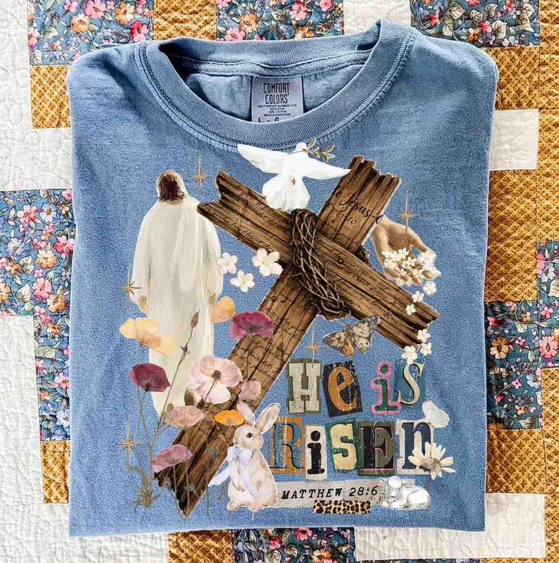 May include: A light blue Comfort Colors t-shirt featuring a graphic design with a cross, dove, flowers, and a bunny. The words "He is Risen" and "Matthew 28:6" are also printed on the shirt.