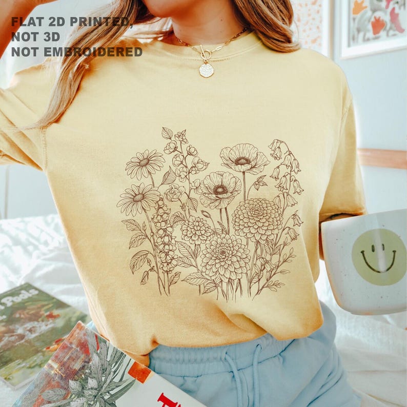 May include: A light yellow t-shirt featuring a detailed brown floral illustration. The text "FLAT 2D PRINTED, NOT 3D, NOT EMBROIDERED" is printed at the top. The shirt is paired with blue shorts and a mug with a smiley face.
