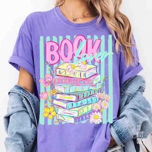 May include: A purple t-shirt with the words "BOOK Lover" in pink and teal. The graphic features a stack of colorful books adorned with flowers, set against a backdrop of vertical teal stripes. The shirt has rolled-up sleeves.