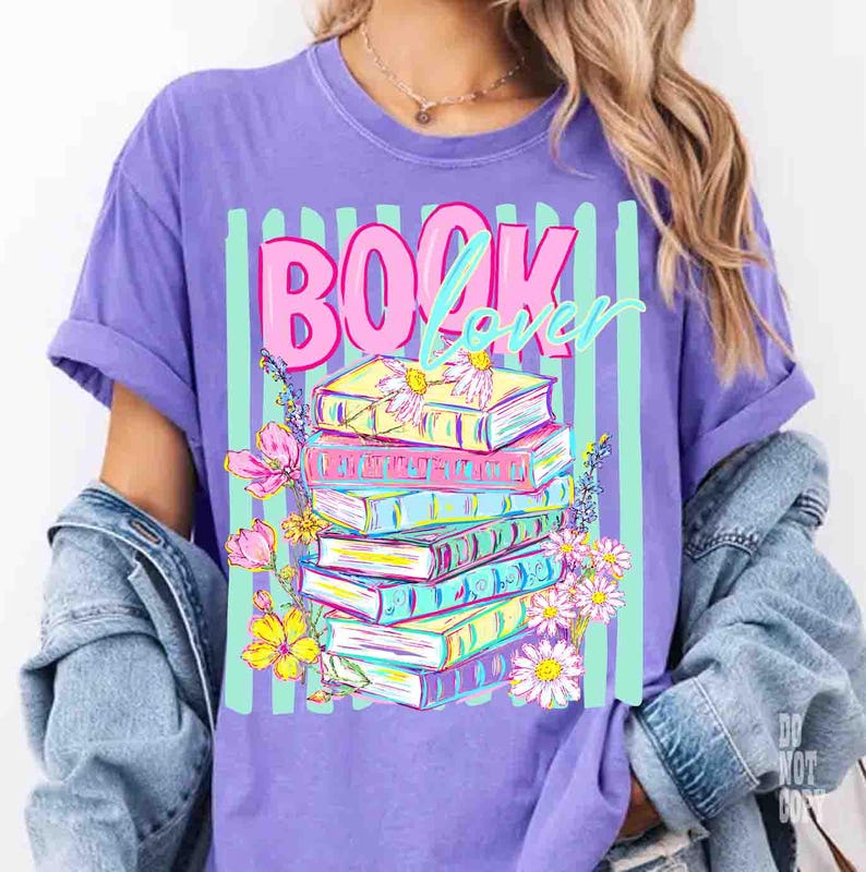 May include: A purple t-shirt with the words "BOOK Lover" in pink and teal. The graphic features a stack of colorful books adorned with flowers, set against a backdrop of vertical teal stripes. The shirt has rolled-up sleeves.