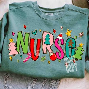 May include: A teal-colored sweatshirt with the word "NURSE" in colorful, festive lettering. The letters are decorated with Christmas-themed graphics, including a Christmas tree, ornaments, and a gingerbread man. The sweatshirt has a crew neck.