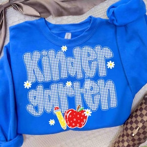 以下が含まれることがあります： 白と青のギンガムチェック柄でKindergartenという文字がプリントされた鮮やかなブルーのクルーネックトレーナー。前面には小さな白いデイジーの花、黄色い鉛筆、白い水玉模様の赤いリンゴのデザインが施されています。