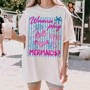 May include: Cream-colored t-shirt with a graphic design featuring the text "Wanna play MERMAIDS?" in pink and blue. The design includes a bikini, starfish, and other aquatic elements. The shirt is a casual, oversized style.