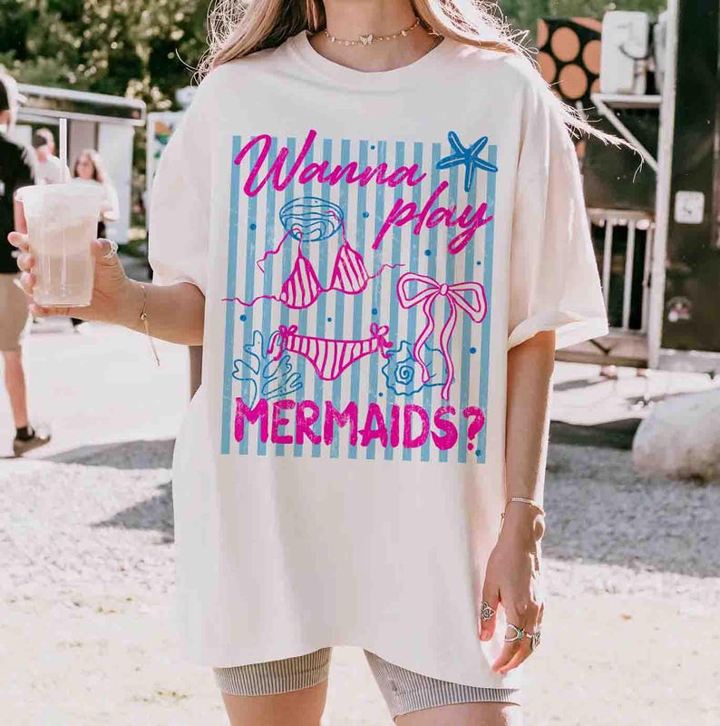 May include: Cream-colored t-shirt with a graphic design featuring the text "Wanna play MERMAIDS?" in pink and blue. The design includes a bikini, starfish, and other aquatic elements. The shirt is a casual, oversized style.