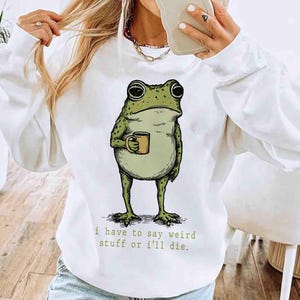 May include: White sweatshirt featuring a whimsical illustration of a green frog holding a mug. The frog has large eyes and a grumpy expression. Below the frog is the text: "i have to say weird stuff or I'll die."