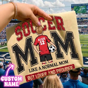 May include: A light tan t-shirt with the words "SOCCER MOM" in red glitter and black glitter. The shirt features a cartoon of a soccer player with the name "ALEX" and the number "12". The text "LIKE A NORMAL MOM BUT LOUDER AND PROUDER" is also on the shirt.