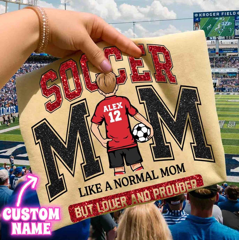 May include: A light tan t-shirt with the words "SOCCER MOM" in red glitter and black glitter. The shirt features a cartoon of a soccer player with the name "ALEX" and the number "12". The text "LIKE A NORMAL MOM BUT LOUDER AND PROUDER" is also on the shirt.