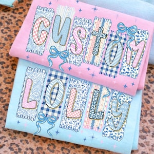 May include: Two folded t-shirts, one pink and one light blue, with the word "CUSTOM" and "LOLLY" printed on them. The letters are decorated with various patterns, including gingham, floral, and stripes, and are accented with blue bows.