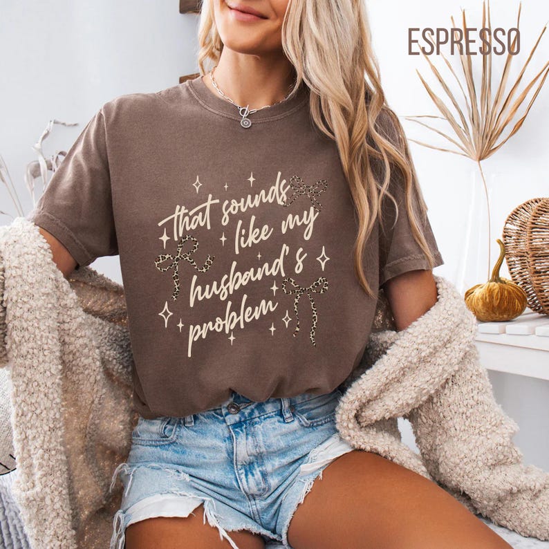 May include: Espresso-colored t-shirt with the text "That sounds like my husband's problem" in a white script font. The shirt features decorative leopard print bows and crosses. The short-sleeved tee is a casual style.