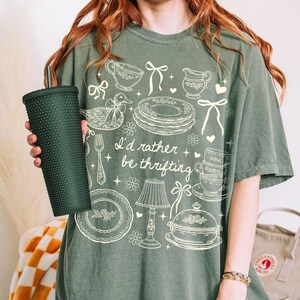 May include: Green t-shirt with the phrase "I'd rather be thrifting" and vintage-style illustrations of teacups, plates, and a lamp. A dark green textured tumbler is held in the image.