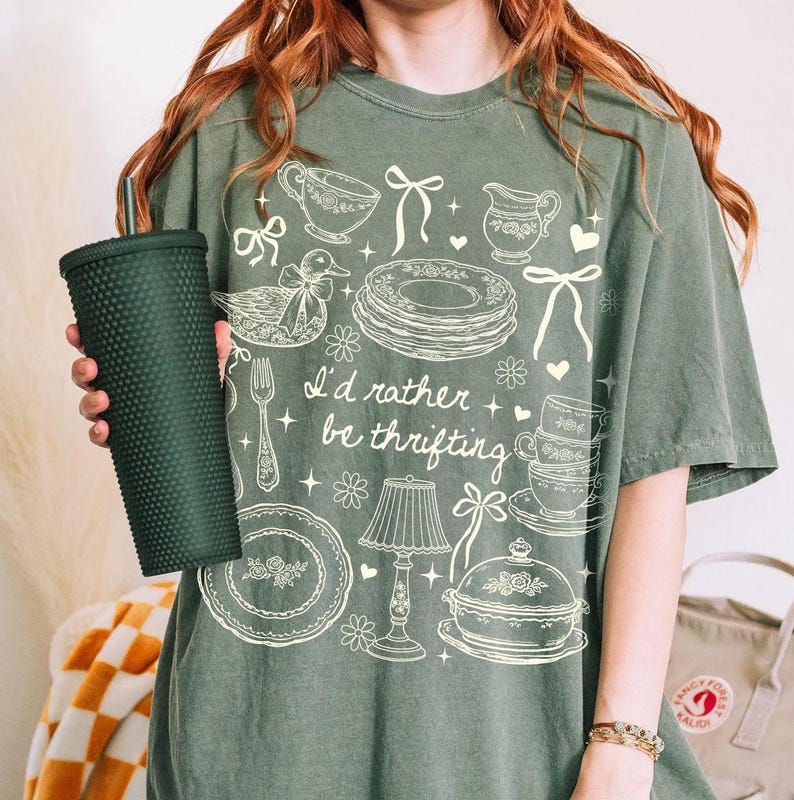 May include: Green t-shirt with the phrase "I'd rather be thrifting" and vintage-style illustrations of teacups, plates, and a lamp. A dark green textured tumbler is held in the image.